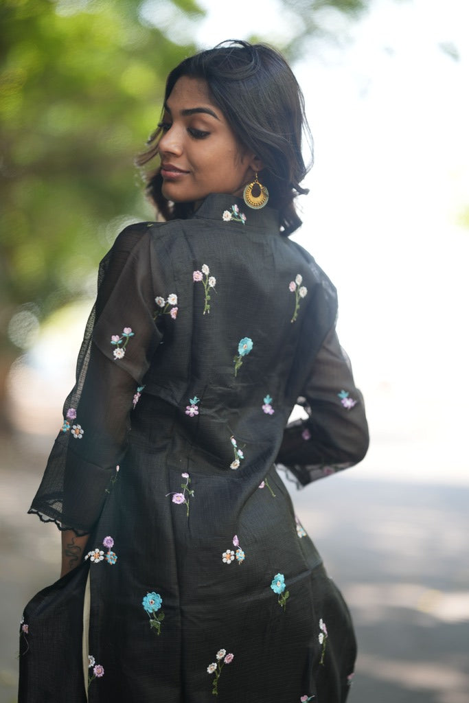 A woman wearing a black sheer embroidered tunic dress with small floral details in blue, pink, and white.